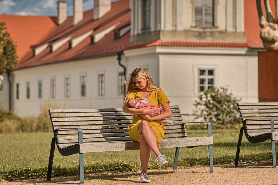 Žena ve žlutých šatech kojí miminko na lavičce v zámeckém parku